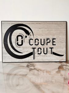 Un panneau rectangulaire sur une surface en marbre affiche le texte COUPE TOUT en noir, avec des lettres stylisées et un dessin circulaire, évoquant la précision de la découpe du jet d'eau sur un fond de pierre claire.