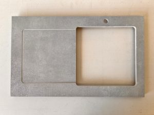 Une plaque rectangulaire, grise, semblable à de la pierre, présente des découpes de jet d'eau : un carré à gauche et un rectangle plus grand surmonté d'un petit trou circulaire à droite, le tout sur un fond uni.