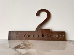 Enseigne en métal comportant le chiffre 2 en haut et les mots chemin de la forge découpés par jet d'eau, présentée sur une surface en marbre sur un fond blanc uni.